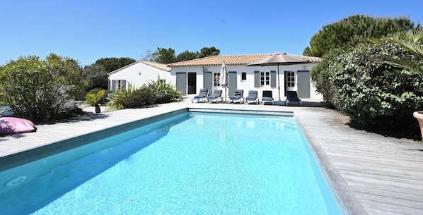A rectangular swimming pool with a wooden deck surrounding it, a white building with shutters, and a clear blue sky.