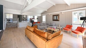 Ein geräumiges Wohnzimmer mit hoher Decke, einem grauen Ecksofa, einem Holztisch und zwei roten Sessel.