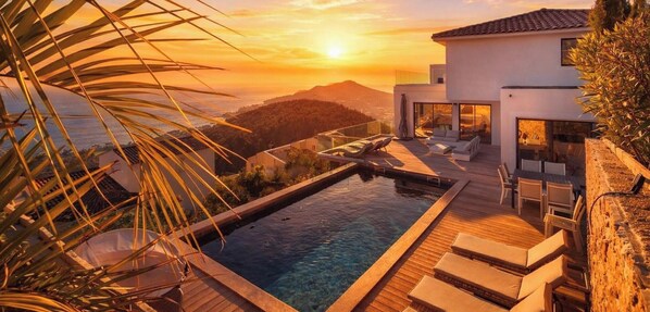 A modern pool with a wooden deck, lounge chairs, and a view of a mountain range at sunset.