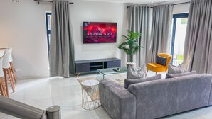 Un salon moderne avec un canapé gris, un téléviseur affichant le message « Bonne Saint-Valentin », une plante en pot et un fauteuil jaune.