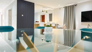 Un espace repas moderne avec une table et des chaises en verre, une cuisine en arrière-plan et un téléviseur fixé au mur.