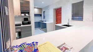 Une cuisine moderne avec des plans de travail blancs, des armoires en bois et un four encastré. On y trouve une fenêtre avec des stores et un évier avec un robinet.
