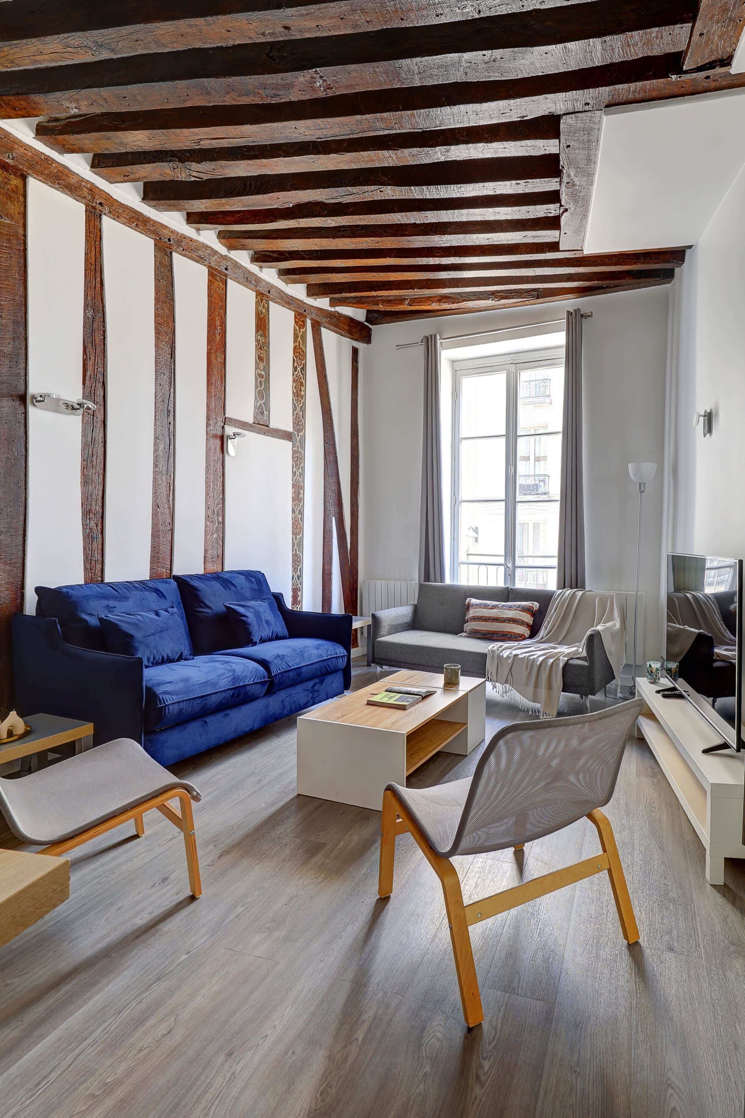 Una sala de estar con vigas de madera a la vista, un sofá azul, un sillón gris y una mesa de centro blanca.