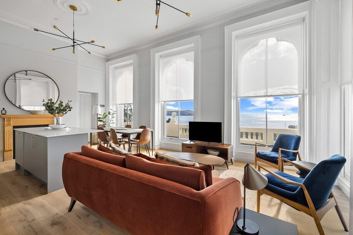 A modern living room with a sofa, chairs, a TV, and large windows offering a view of the sea.