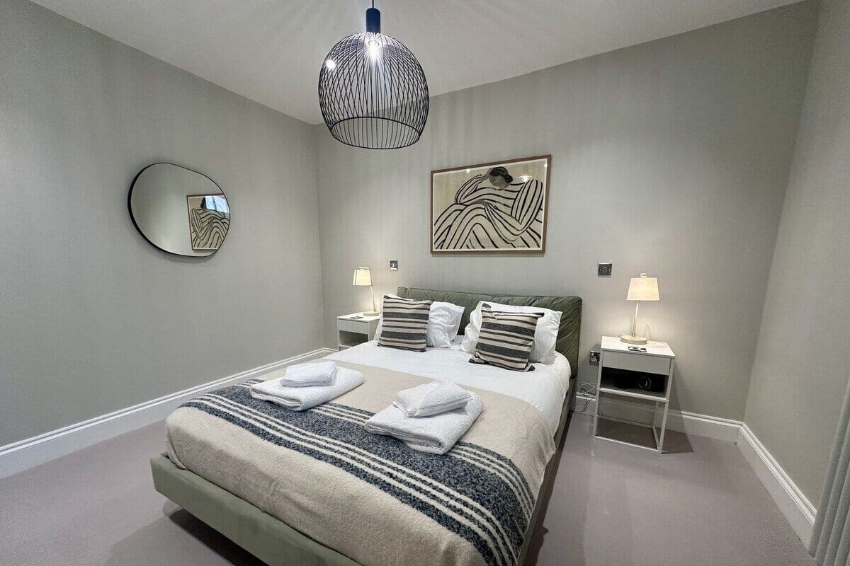 A neatly arranged bedroom with a large bed, two bedside tables, a wall-mounted mirror, and a hanging light fixture.