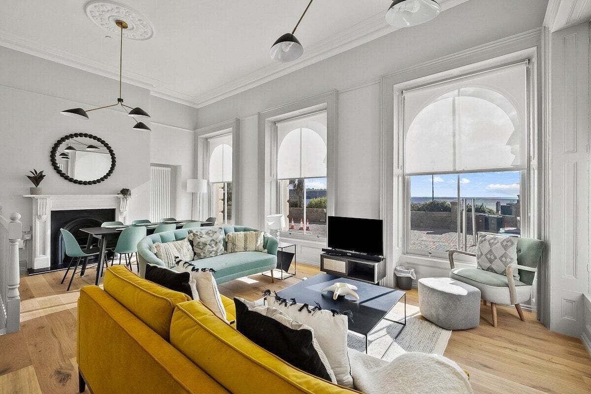 A spacious living room with a yellow sofa, teal and white armchairs, a flat-screen TV, and large windows offering a view of the outdoors.