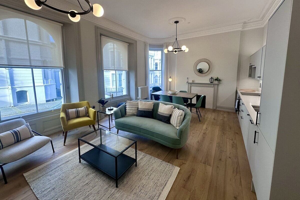 A modern living room with a sofa, armchairs, and a coffee table. There is a dining area with a table and chairs. The room has large windows and a view of the city.