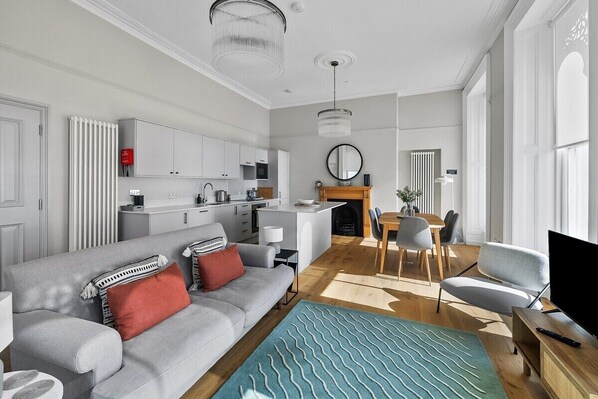Una sala moderna con un sofá gris, almohadas rojas y blancas, un tapete azul estampado, una mesa de comedor de madera con sillas, una zona de cocina con armarios blancos y un espejo grande en la pared.