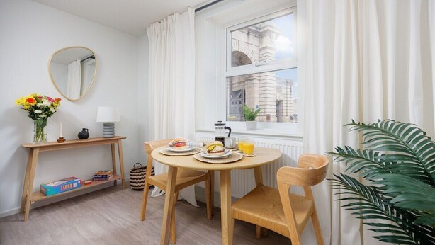 A dining area with a round table, two chairs, a mirror, a shelf with books, a vase with flowers, and a window with curtains. - Luxury Ground Floor 2 bed Apartment (Plymouth)