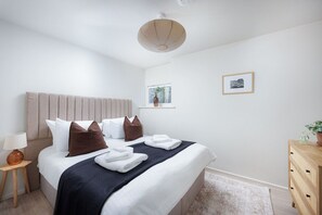 A neatly made bed with pillows, a bedside table with a lamp, a wooden dresser, and a framed picture on the wall. - Luxury Ground Floor 2 bed Apartment (Plymouth)
