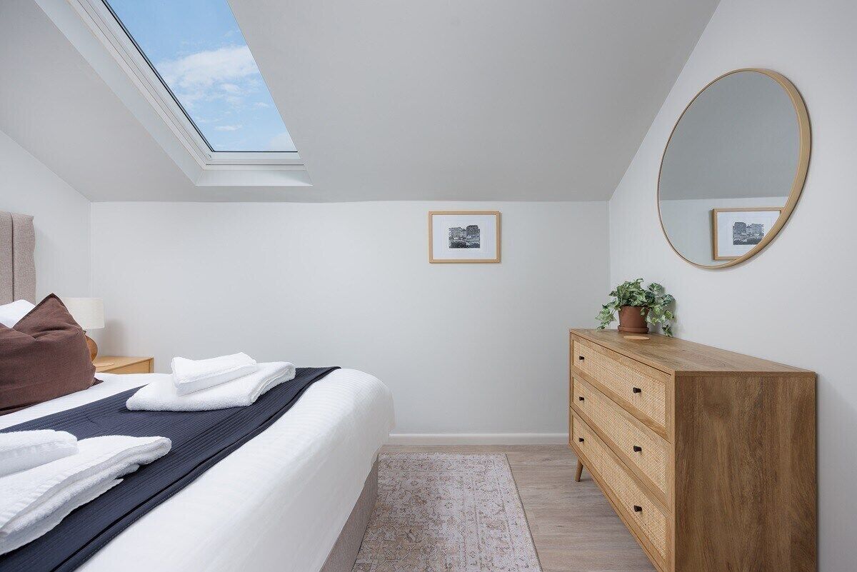 Ein Schlafzimmer mit Dachfenster, einem Bett mit weißen Leinentüchern, einer Holzkommode und einem gerahmten Bild an der Wand.
