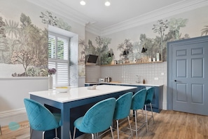 A modern kitchen with a central island, bar stools, a window with shutters, and a door. - Windsor Villas by Pureserviced (Plymouth)