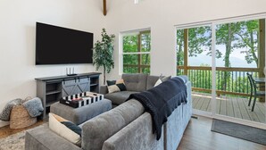Ein modernes Wohnzimmer mit einem großen Flachbildfernseher, grauen Sofas und einem Holzboden mit Blick auf Bäume.