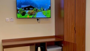 A wooden desk with a television mounted above it, a small refrigerator under the desk, and a wooden wardrobe to the right. - SUNSET HOTEL (Al Amarat)