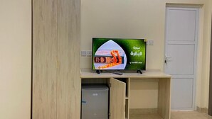 A hotel room with a bed, a TV on a wooden stand, a small fridge, and a door. - SUNSET HOTEL (Al Amarat)