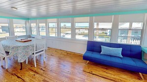 Um quarto com piso de madeira, uma mesa com cadeiras, um sofá azul e janelas grandes com vista para a praia.