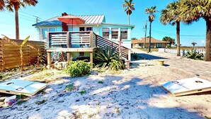 Uma casa na praia com varanda, palmeiras e chão de areia.