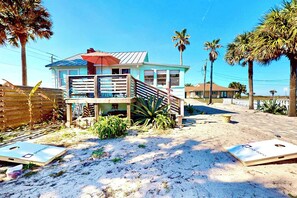 A beachfront house with a balcony, palm trees, and a sandy ground.