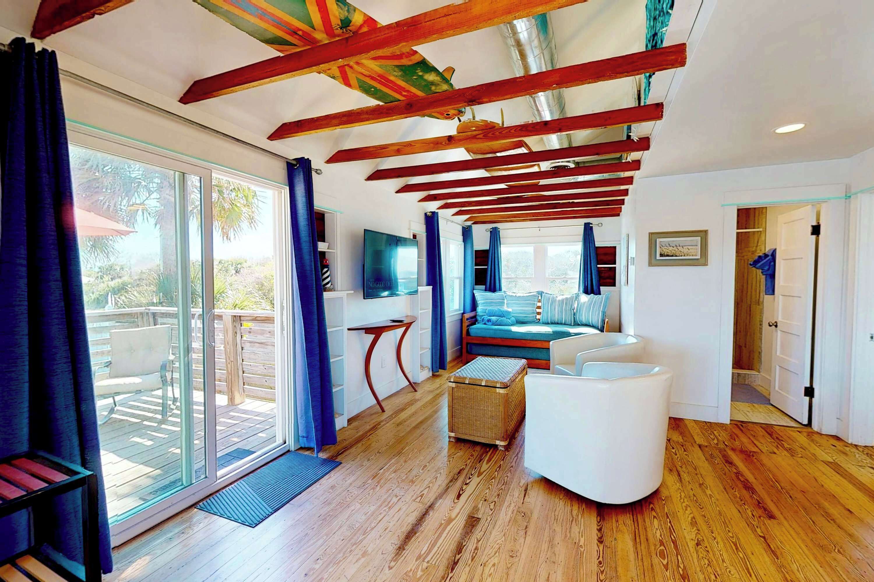 A modern interior with a wooden ceiling, a seating area with a sofa and ottoman, a television, and a sliding glass door leading to an outdoor deck.