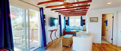 A modern interior with a wooden ceiling, a seating area with a sofa and ottoman, a television, and a sliding glass door leading to an outdoor deck.