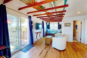 A modern interior with a wooden ceiling, a seating area with a sofa and ottoman, a television, and a sliding glass door leading to an outdoor deck. - Coastal Cottage, Sandy Backyard & Steps to Beach (St. Augustine)