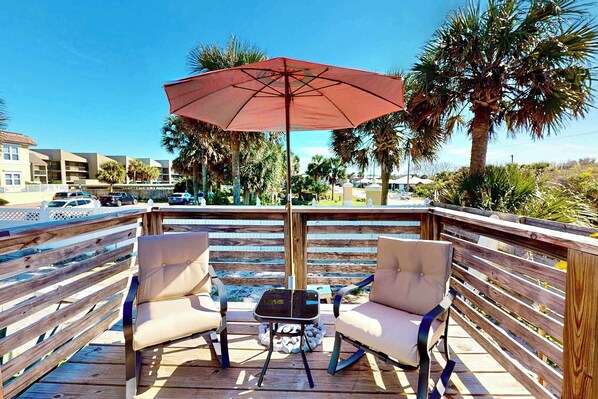 An outdoor deck with two cushioned chairs and a table under a red umbrella, overlooking a parking area and buildings.