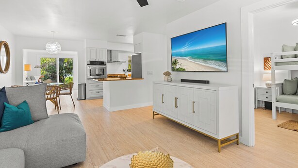 A modern living room with a grey sofa, a dining area, a kitchen, and a large TV displaying a beach scene. - 3-bedrooms-2 Full Baths,  Private backyard, mnts to Central Beach & downtown (Vero Beach)
