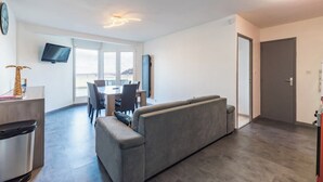 Una sala de estar moderna con un sofá gris, una mesa de comedor con sillas, un televisor de pantalla plana montado en la pared y un gran ventanal con vistas a la ciudad.