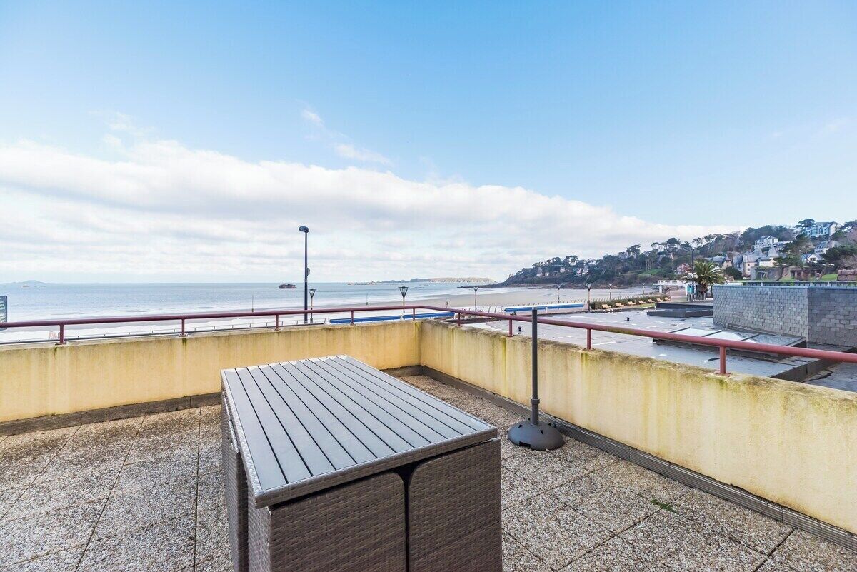 Une terrasse sur le toit, avec une table en bois, une rambarde en métal et une vue sur la mer et une ville située sur une colline.