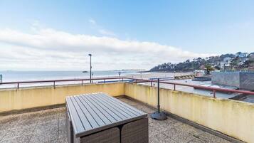 Une terrasse sur le toit, avec une table en bois, une rambarde en métal et une vue sur la mer et une ville située sur une colline.