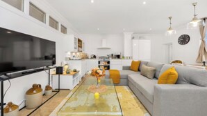 Un salon moderne avec un canapé gris, une table basse et un téléviseur. On aperçoit à l’arrière-plan un coin cuisine avec des meubles blancs et un four à micro-ondes.