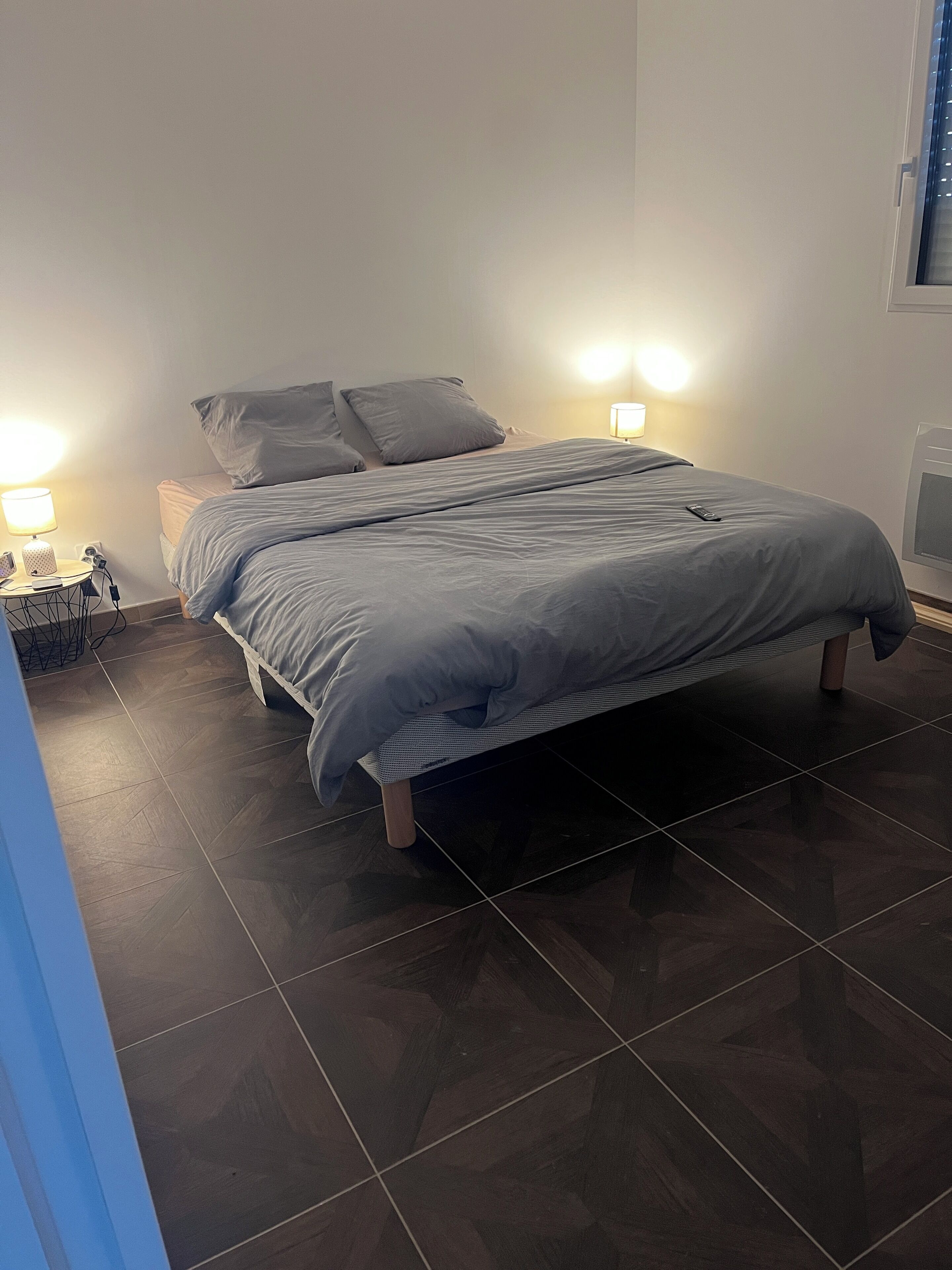 Een net opgemaakt bed met een grijze dekbedovertrek en kussens, een nachtkastje met een lamp, en een wandradiator.