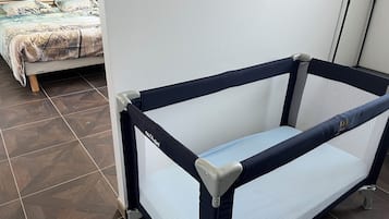 A baby crib with a mesh base and a padded mattress, positioned against a white wall in a room with a tiled floor.