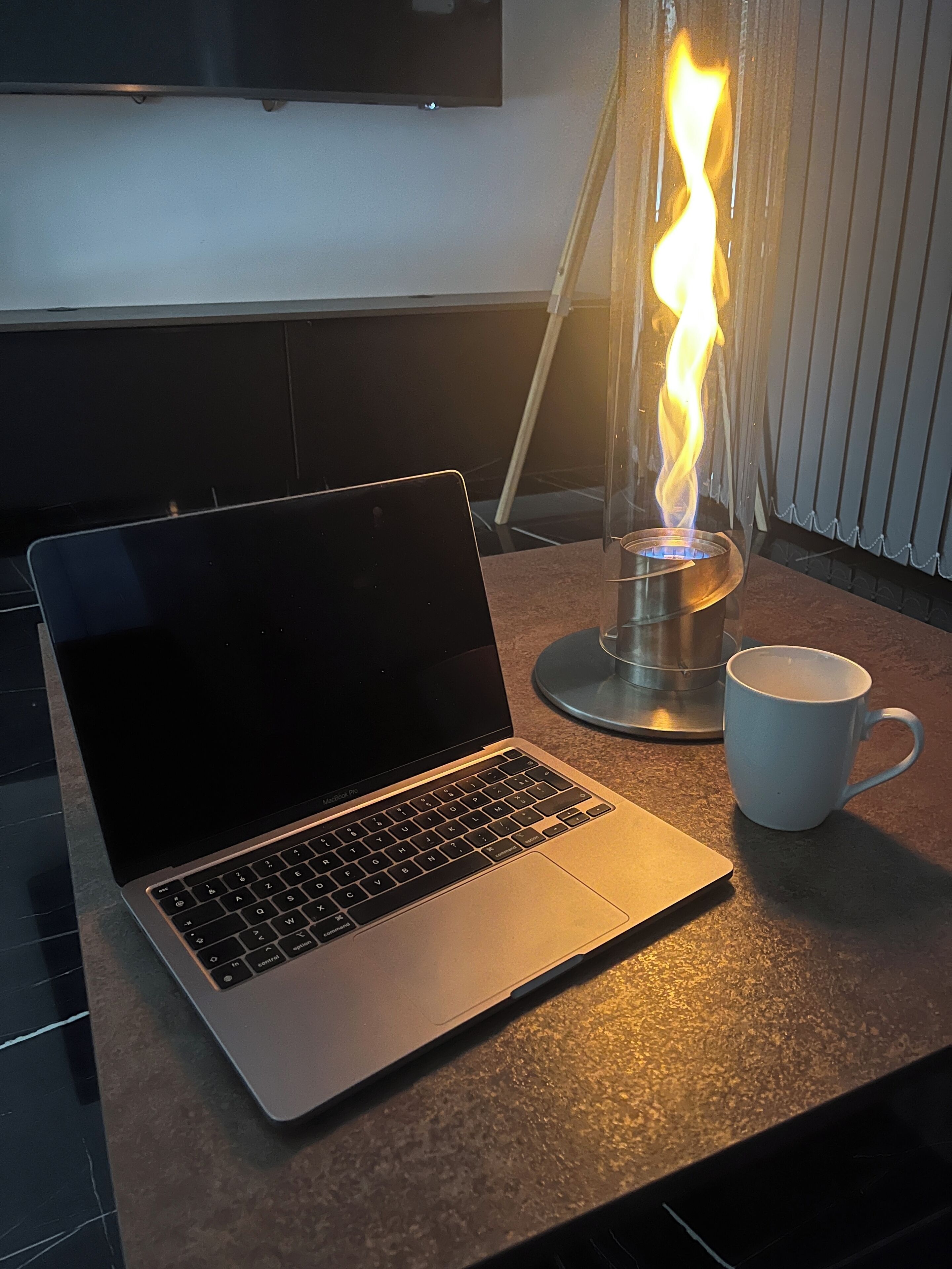 Um laptop sobre uma bancada, com um recipiente de vidro transparente com fogo aceso, uma caneca branca e uma televisão de tela plana fixada na parede.