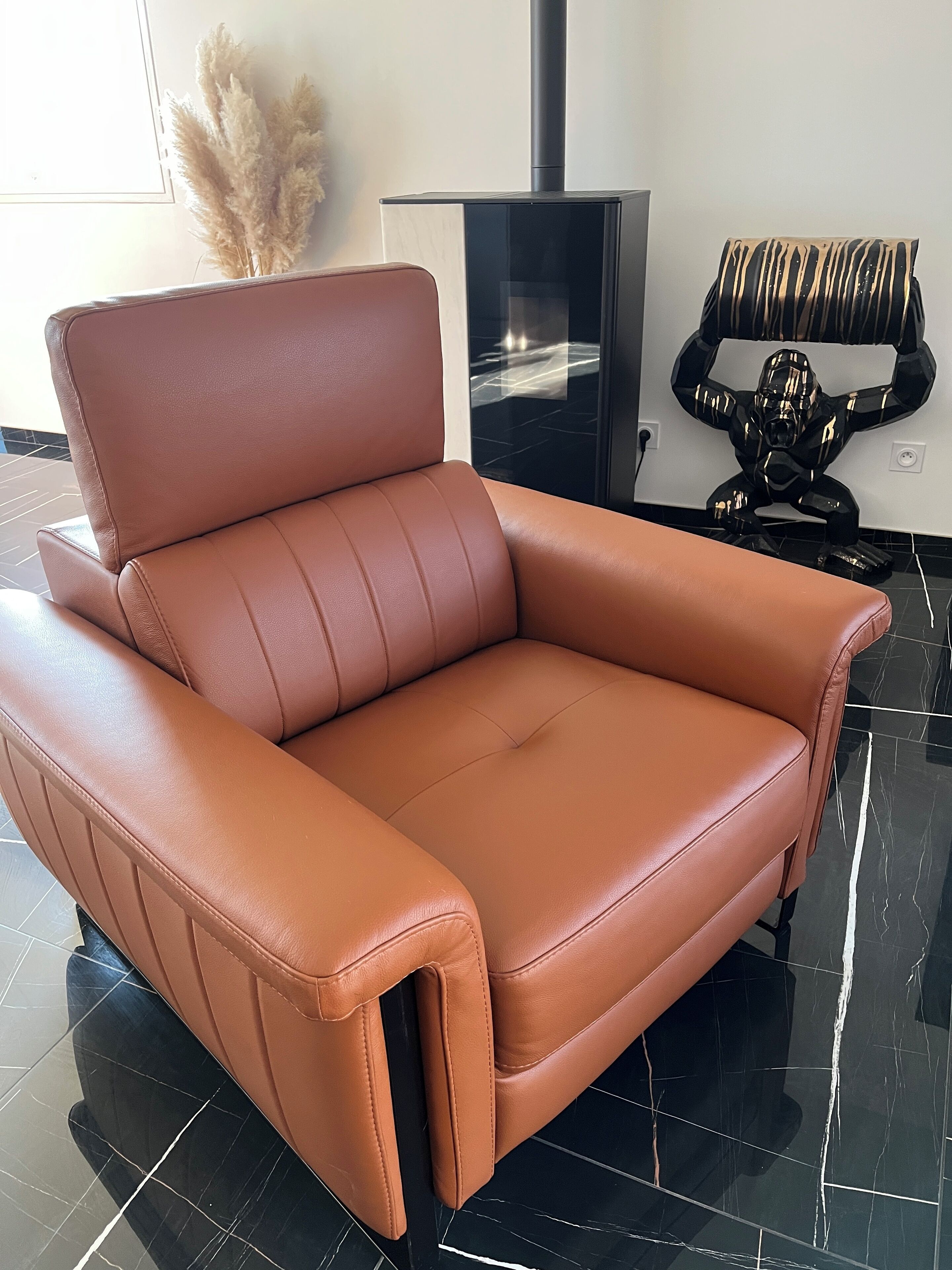 Een bruine leren bank met een hoge rugleuning en armleuningen, geplaatst in een kamer met zwart betegelde vloeren en een zwarte open haard.