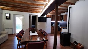 A dining area with a long wooden table, red chairs, and a central bowl. There is a kitchen area with a stove and a TV mounted on the wall.