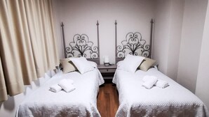 A room with two beds, each with a white bedspread and pillows, a nightstand with a lamp, and a window with curtains.