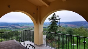 A balcony with a wooden table and chairs, offering a view of a green landscape with trees and hills.