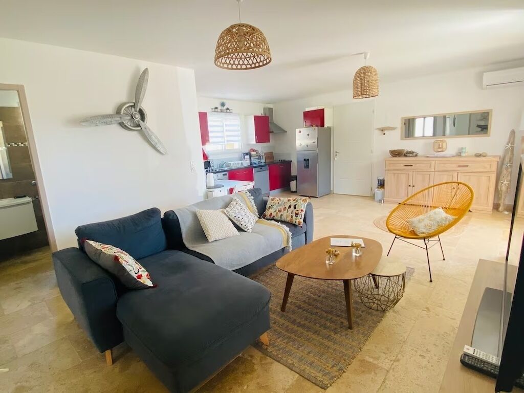 A modern living room with a sofa, coffee table, and a wicker chair. A kitchen with red cabinets and a dining area with a table and chairs are visible in the background.