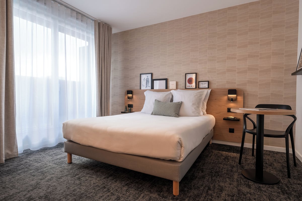 Habitación de hotel moderna con una cama grande, una mesita pequeña y un escritorio. Hay cuadros en la pared y una ventana con cortinas.