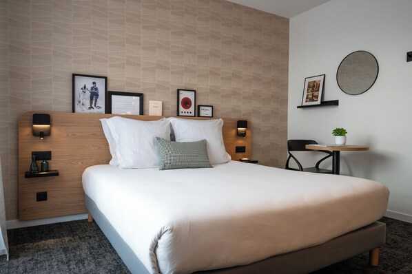 Una cama bien hecha con almohadas blancas y a cuadros, cabecera de madera, lámparas de noche, una mesita con una planta y cuadros en la pared.