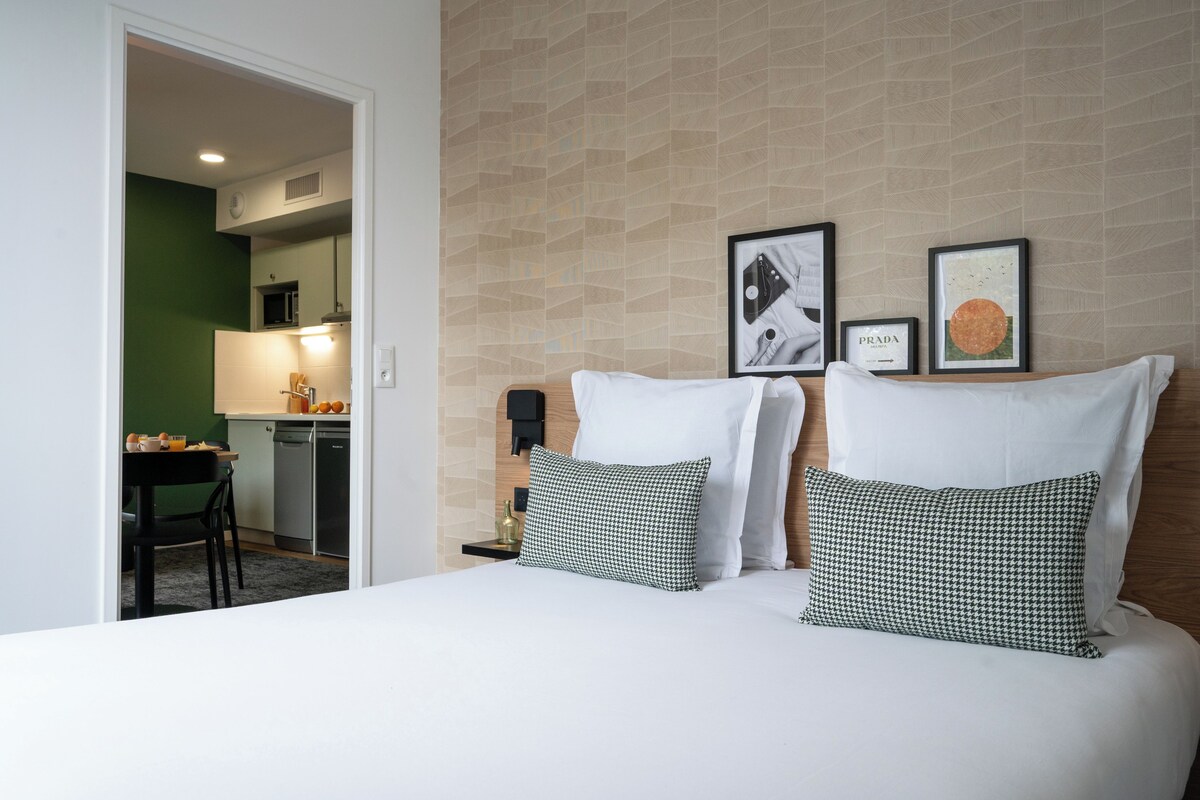 A hotel room with a bed, two pillows, a nightstand, a wall-mounted clock, and framed pictures on the wall.