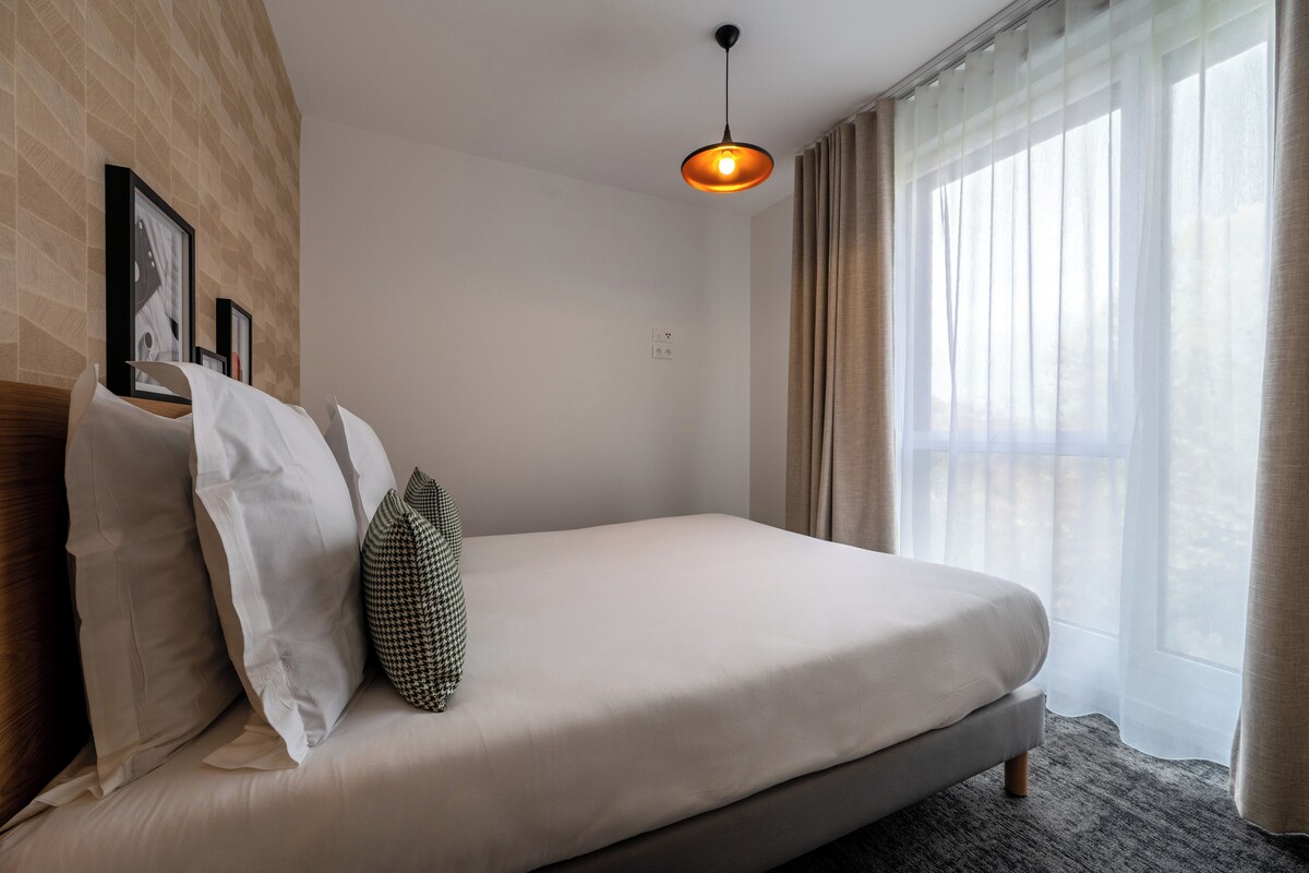 Un llit ben fet amb matalàs de color clar, una sola almoçada i una almoçada decorativa. Hi ha un llum penjant sobre el llit. L'habitació té una finestra gran amb cortines primes que deixen entrar llum natural.