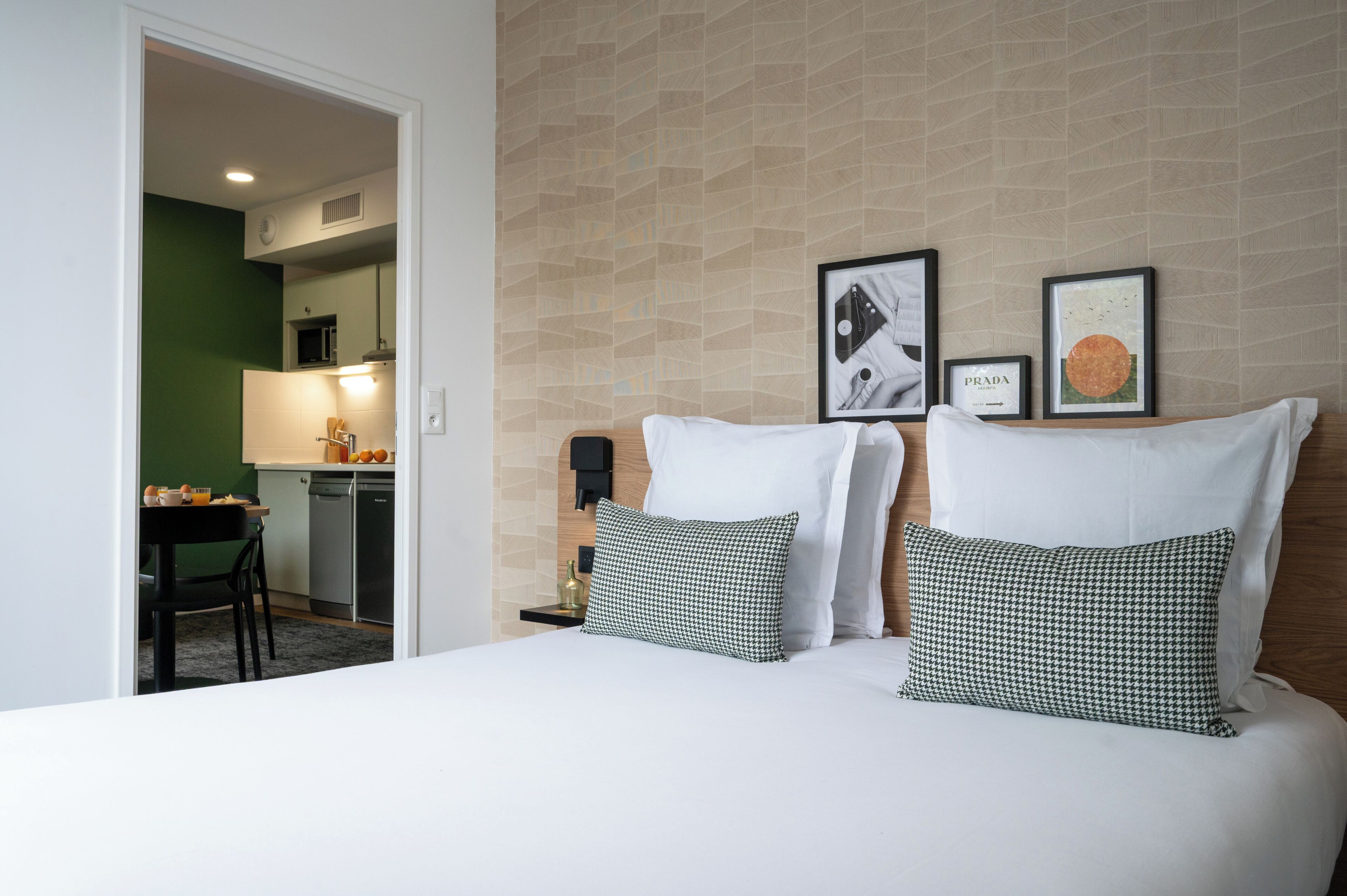 A hotel room with a bed, two pillows, a nightstand, a framed picture, a wall clock, and a view into the kitchen area.