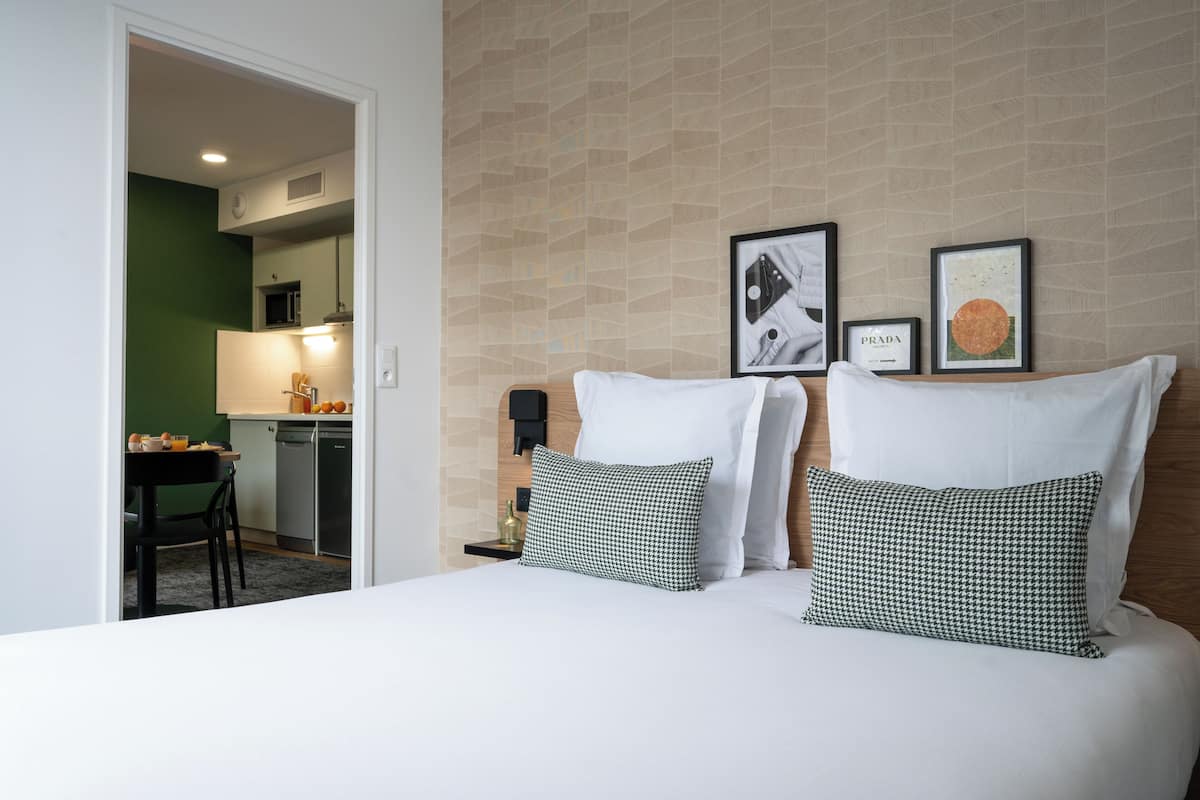 A hotel room with a bed, two pillows, a nightstand, a framed picture, a wall clock, and a view into the kitchen area.