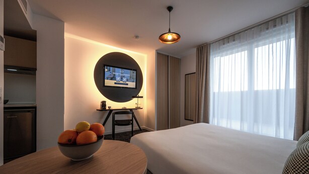 Habitación de hotel con cama, escritorio con monitor de computadora, frutero y ventana con cortinas.