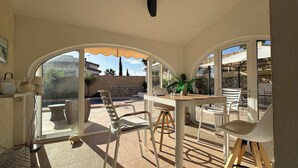 Un patio iluminado por el sol con una mesa y sillas, un muro de piedra y vistas a una piscina y edificios al fondo.