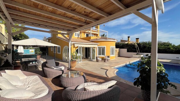 Un espacio al aire libre cubierto con mobiliario de mimbre, una piscina y un edificio amarillo al fondo.