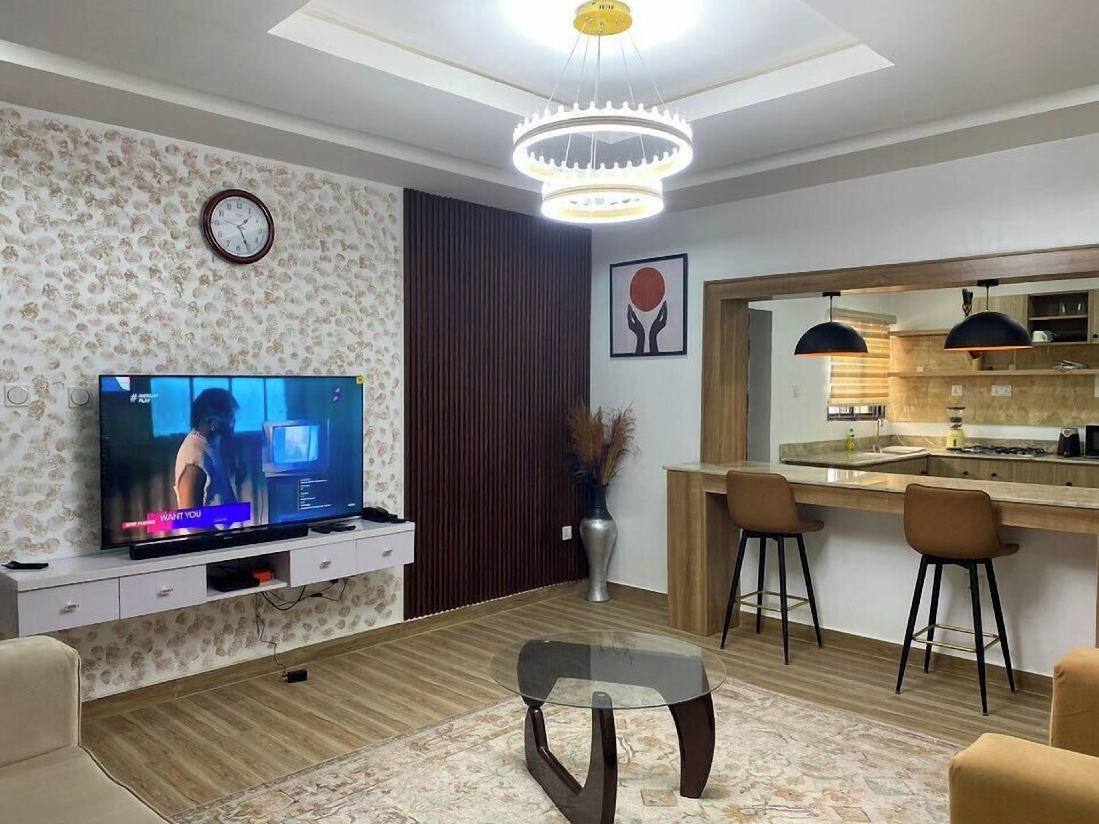 Una sala moderna con un televisor de pantalla plana, un comedor con una cocina al fondo y un reloj de pared decorativo.
