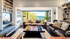 A spacious living room with a large window overlooking the sea, a chessboard on the coffee table, and a television on the left. - 3bed Apt 4-min to Llandudno Beach Backline Views (Cape Town)